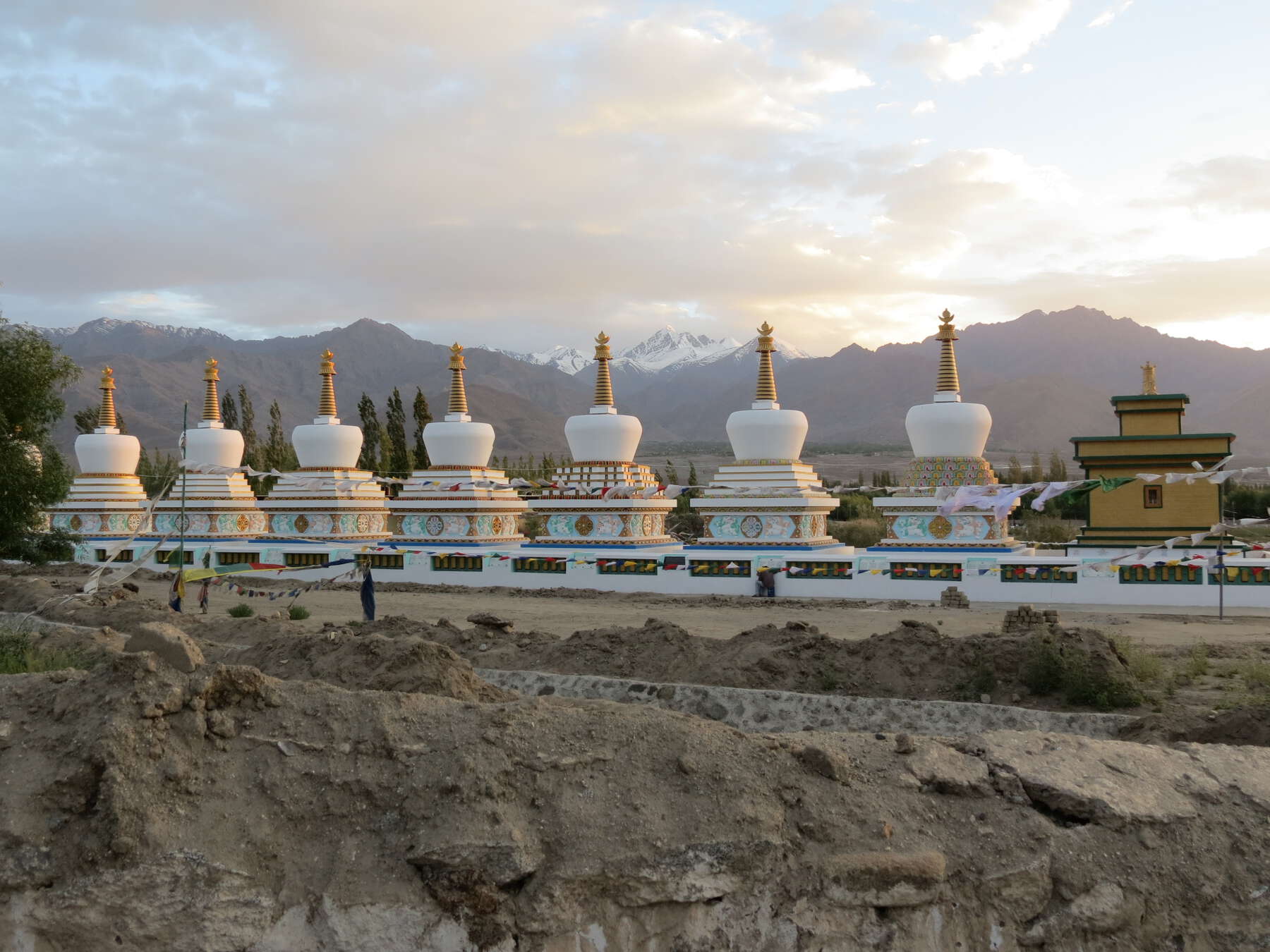 Seven decorated stupas and a small square building lined up consecutively, with a scenic view of a mountain ridge at dawn.