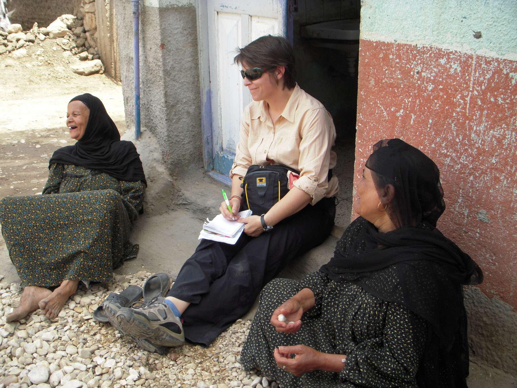 Three women smiling, the one in the middle is taking notes, while they all are sitting on the pebbled ground near a doorway.