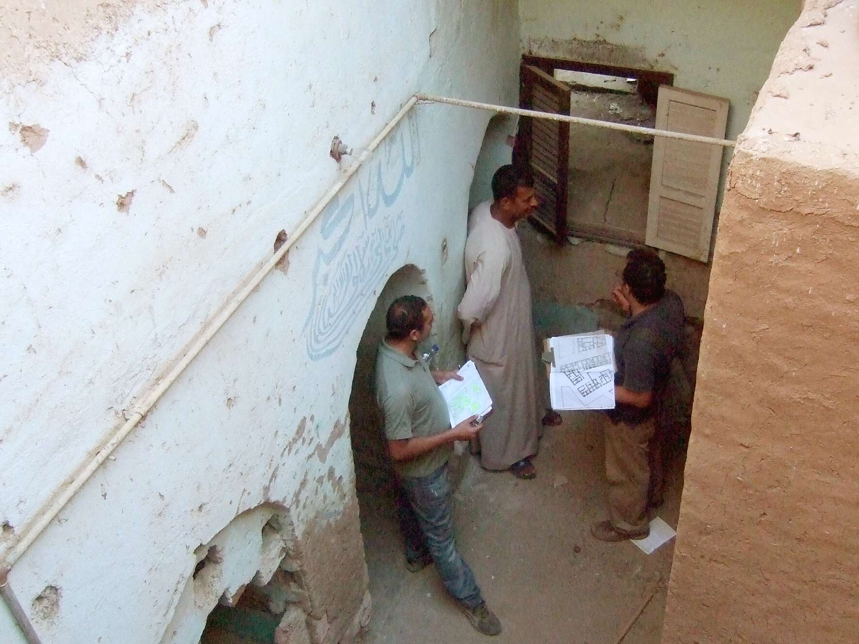 Elevated view of three men talking at the end of a hallway next to a window, two of them are holding building layouts.