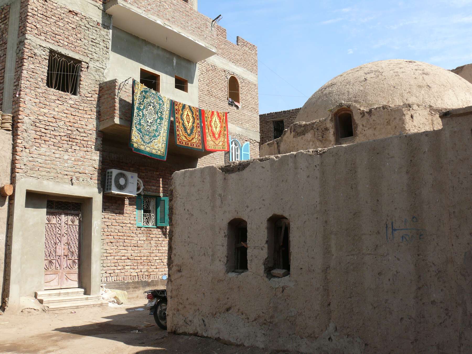 Small building with a dome on the top and behind it, a multistory brick building with three colorful rugs hanging from a balcony.