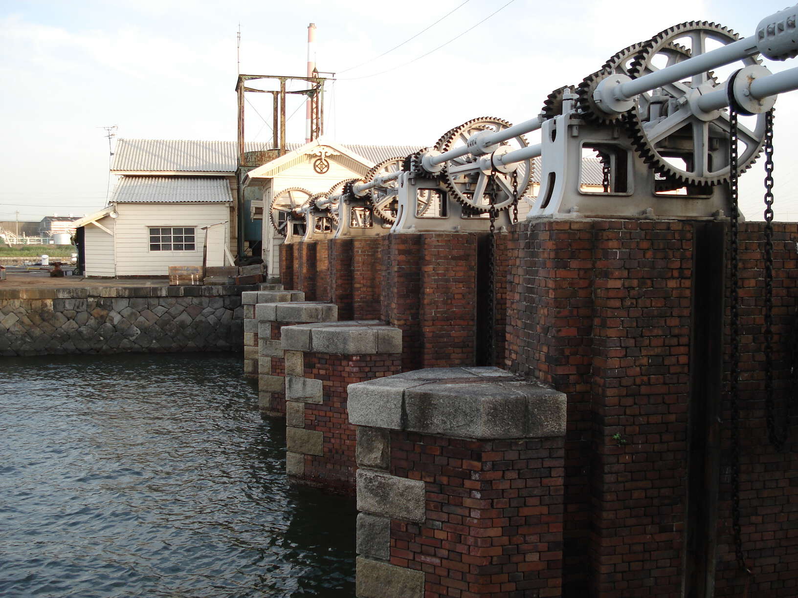 Metal sluice gate system on top of a tall, brick wall blocking off ocean water, and a building next to it in the background.