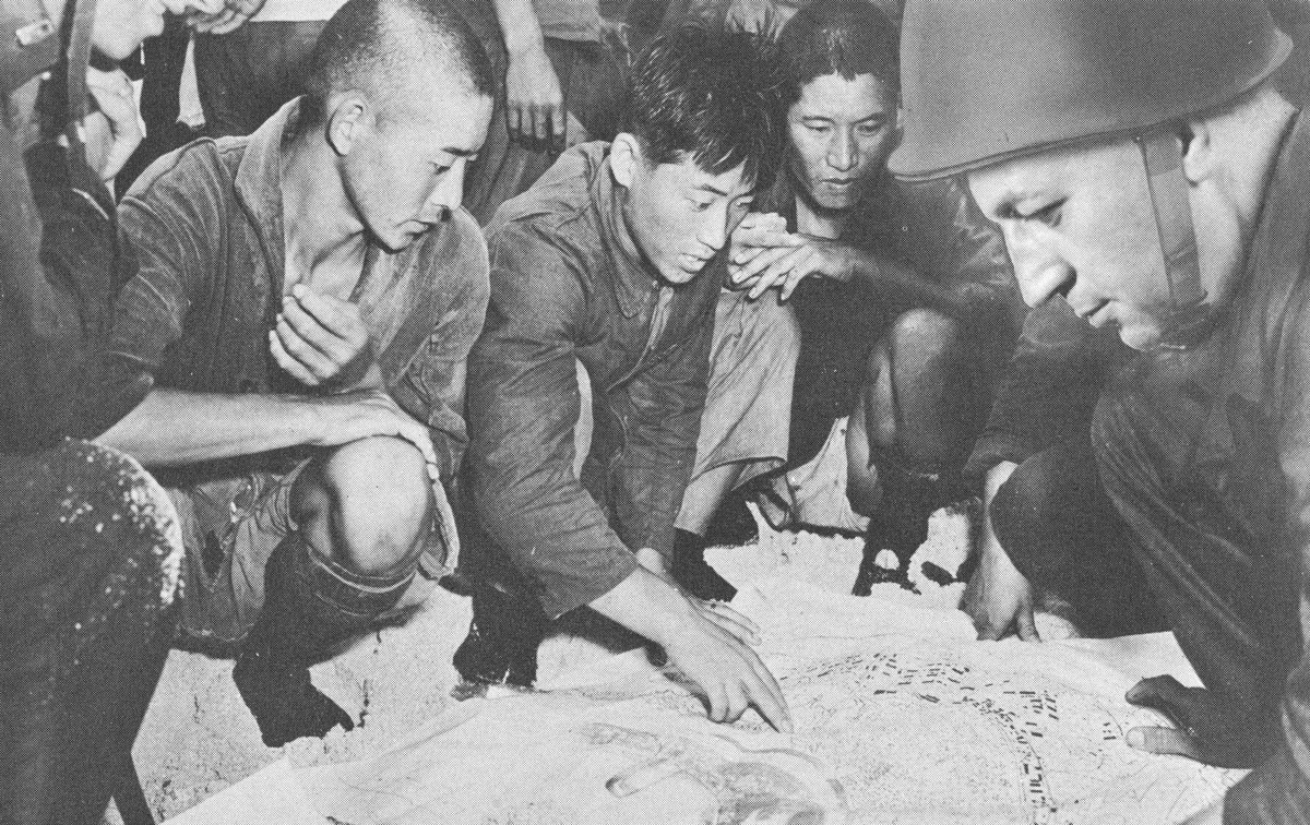 Group of men crouching down in front of a large map. A Korean laborer points to a part of the map while a soldier looks on.