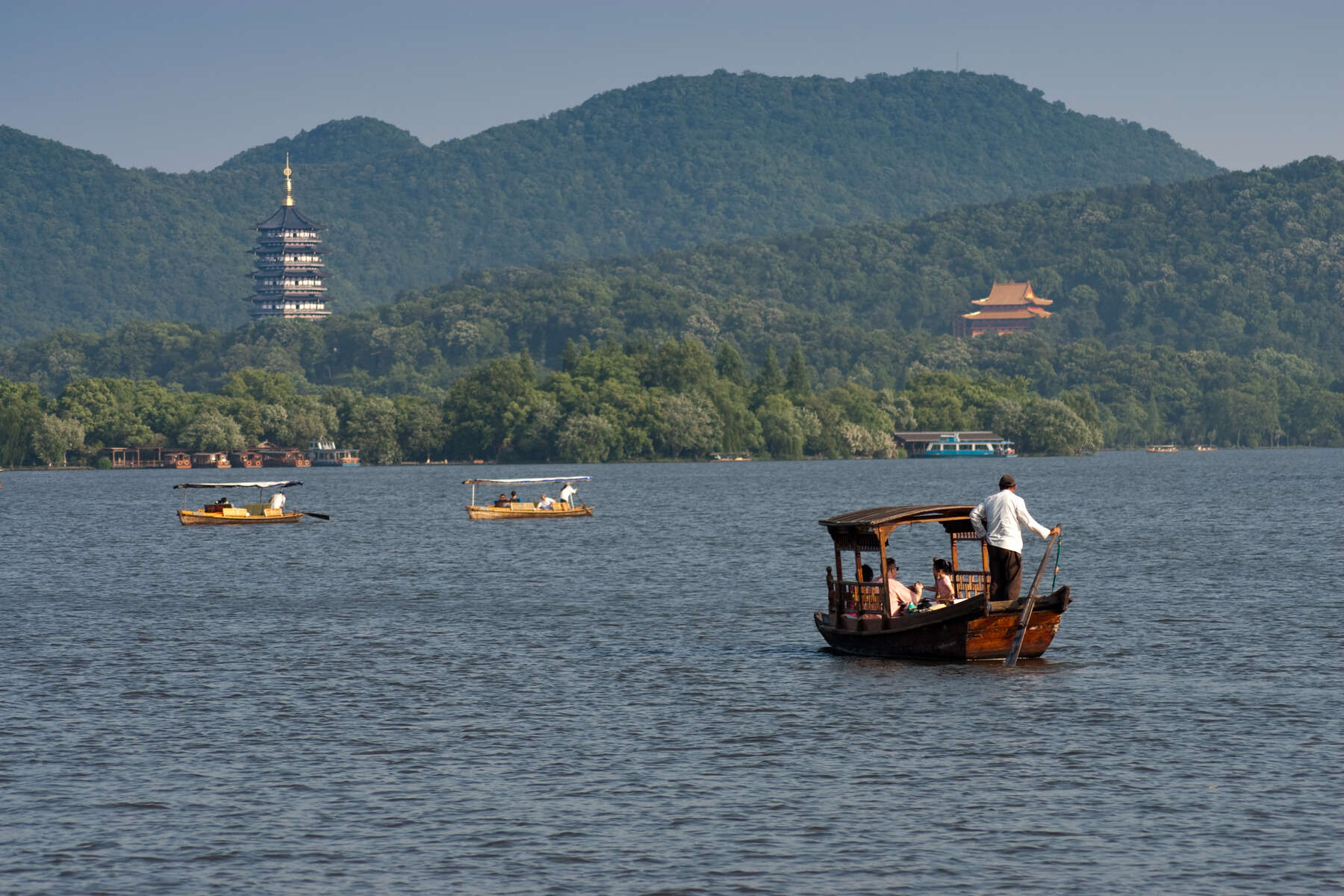 Four boats sailing in a lake. In the distance, two multistory structures standout on a mountain completely covered in trees.