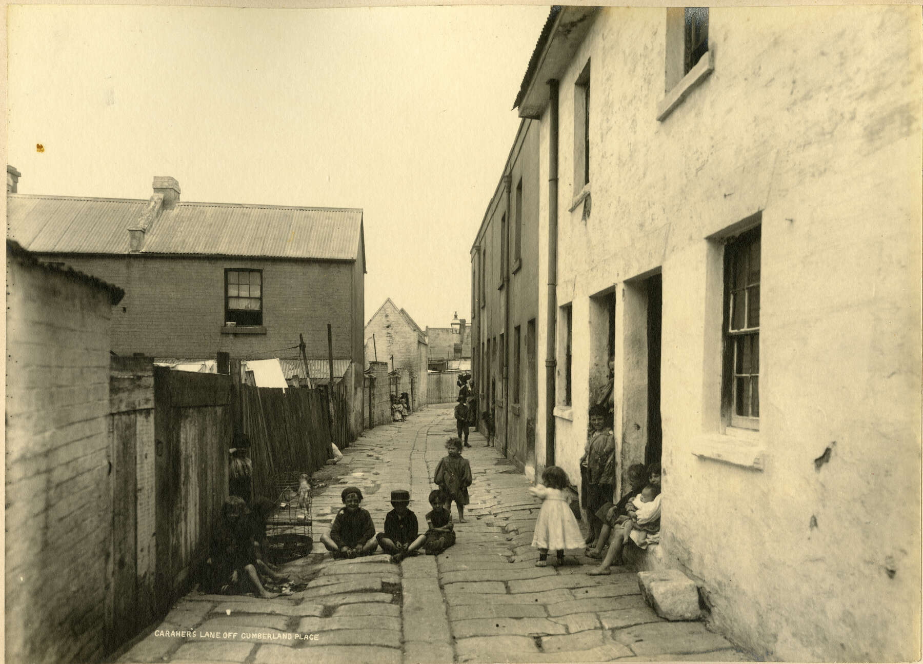 Old sepia photograph of a small lane with houses on each side and children hanging out on the walkway.