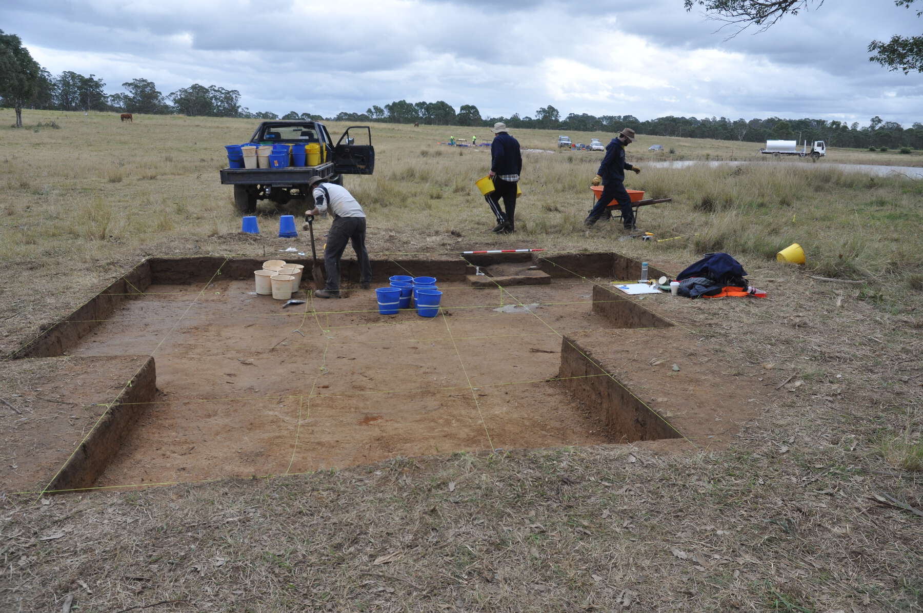 Three men work on an excavation in an open field. In the back, a truck is loaded with several buckets of their findings.