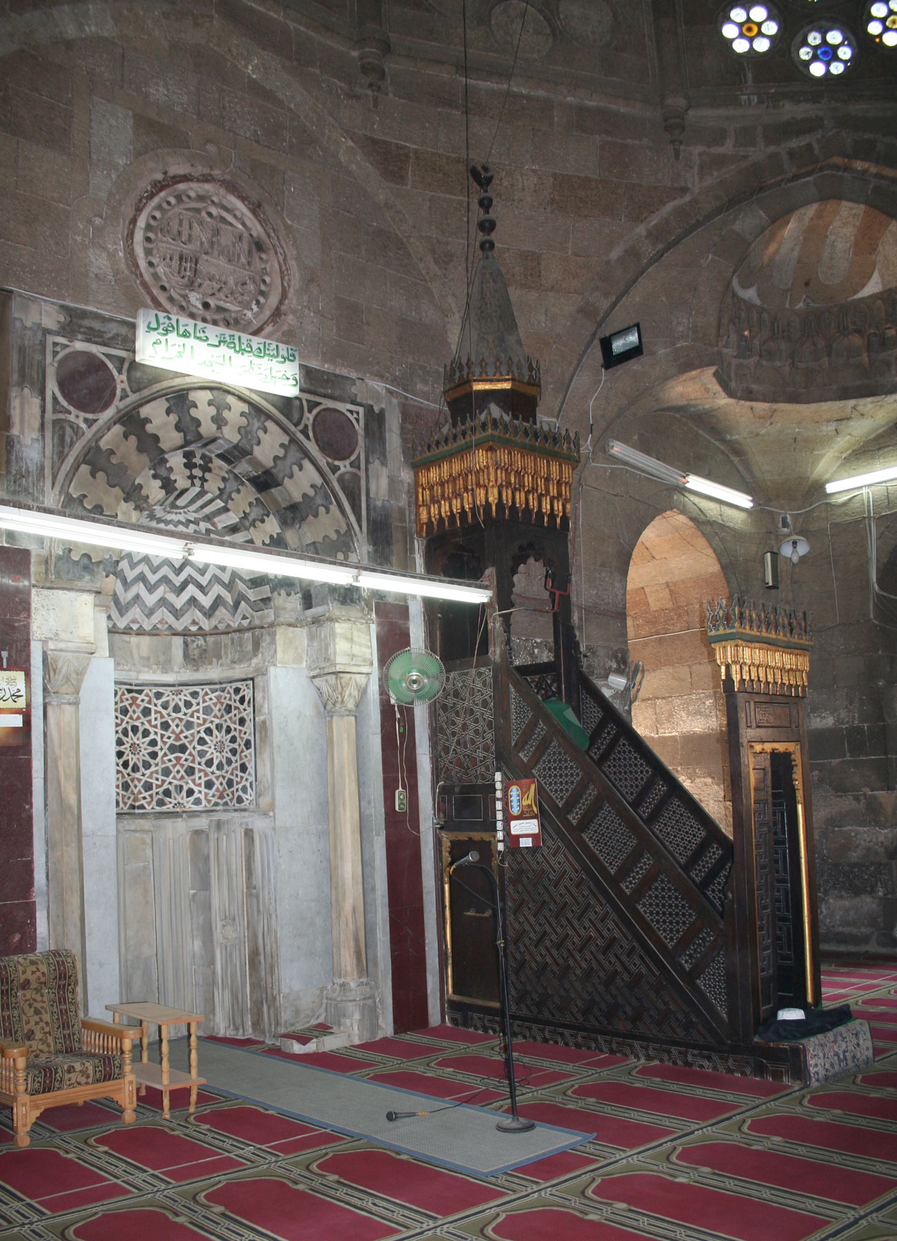 Fans and several light fixtures are attached to the delicate walls of a mosque. At the center is a microphone stand.
