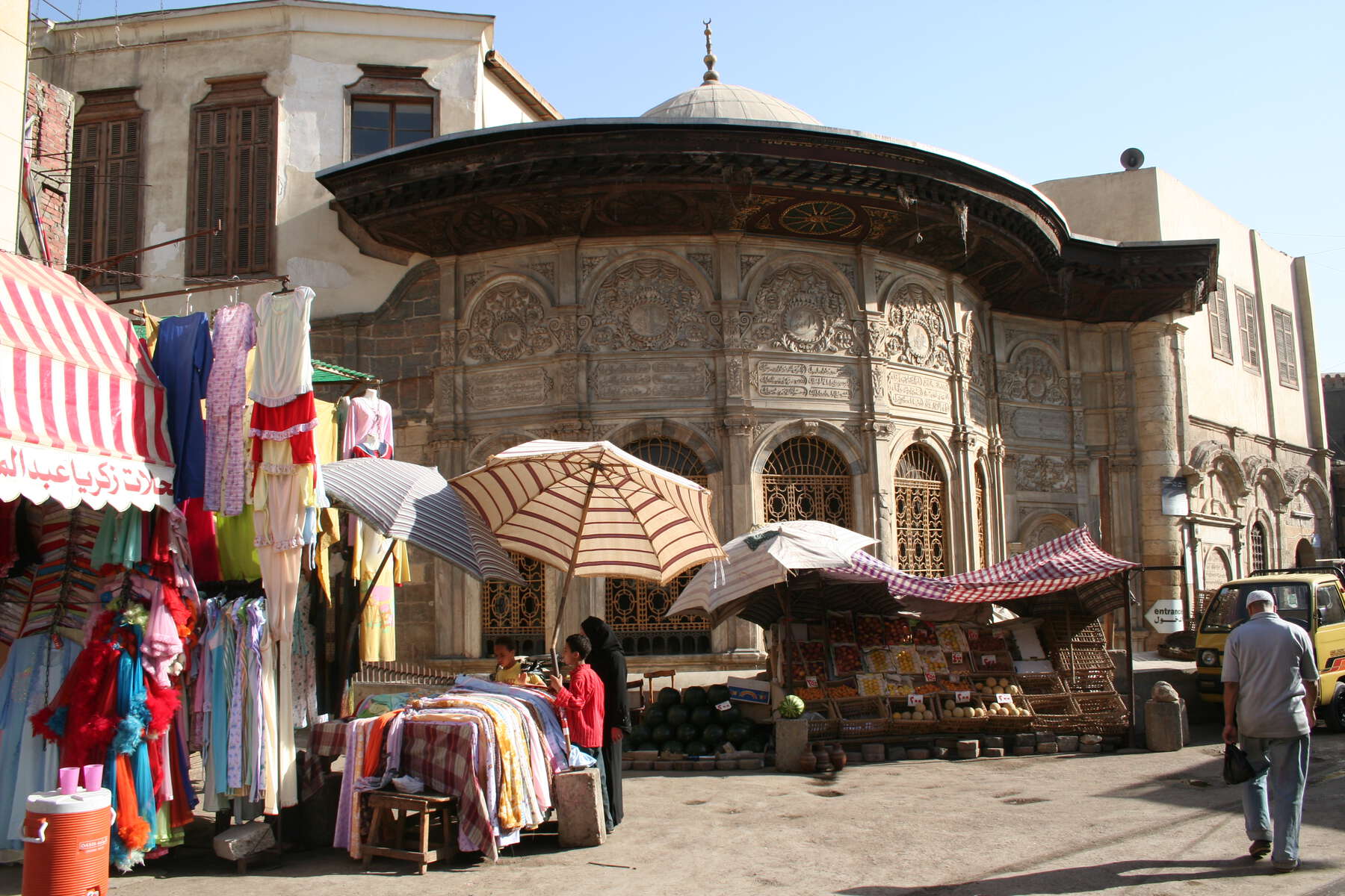 Street market selling food and clothes in front of a dome-shaped sabil covered with ornamentation and arches at the bottom.